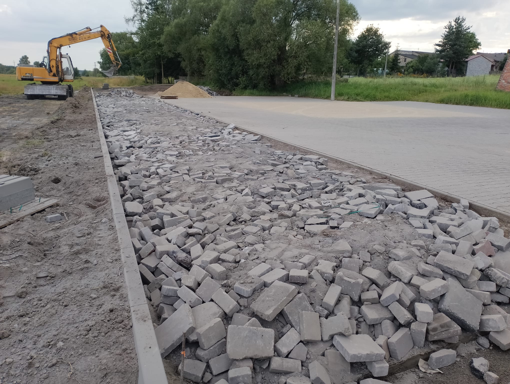 Rozszerzanie utwardzenia terenu w miejscu organizacji Jarmarku nad Liswartą. Widoczny ułożony krawężnik i wstępnie rozłożony gruz, a w tle także nawierzchnia wybrukowana kostką oraz pracująca koparko-ładowarka.