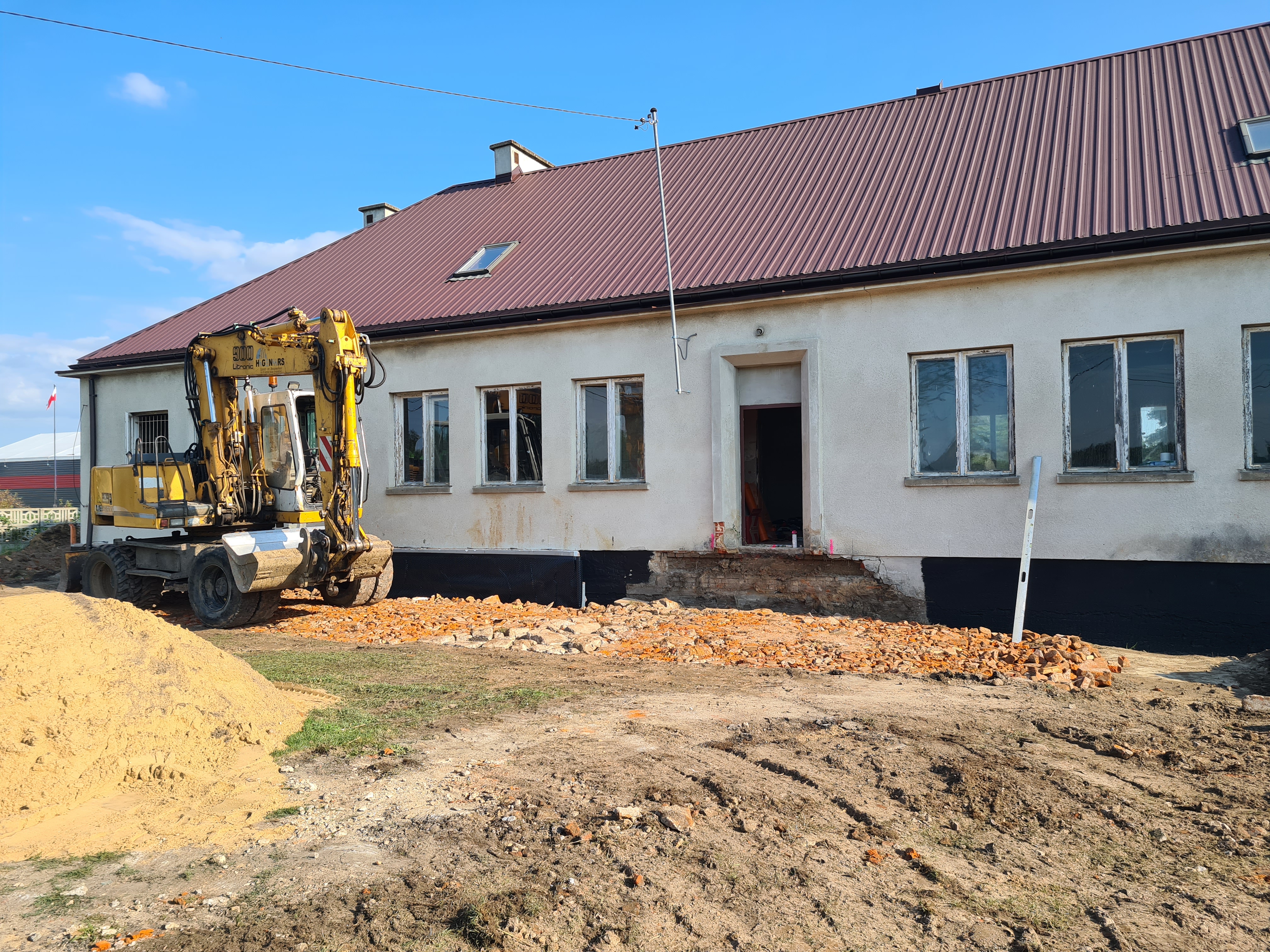 Prace remontowe przy budynku świetlicy w Grabarzach. Widoczna elewacja budynku i miejsce po zdemontowanych starych schodach wejściowych oraz teren utwardzony gruzem ceglanym. Obok po lewej pryzma piasku, a za nią koparko-ładowarka.