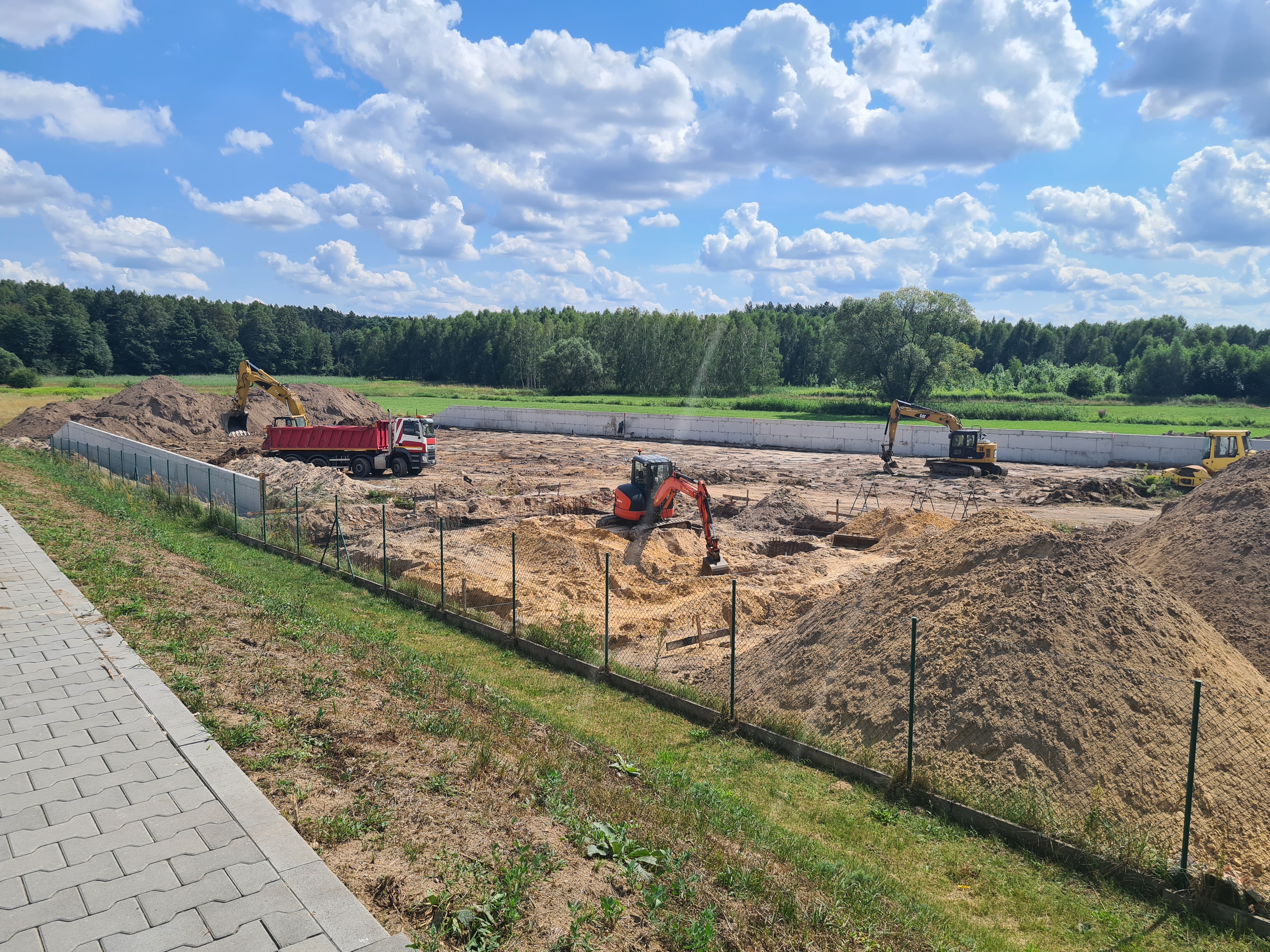 Widok ogólny terenu realizacji budowy PSZOK w Lipiu. Wewnątrz ogrodzenia z siatki znajdują się pryzmy piasku i ziemi, przy wykopach pracują koparko-ładowarki, a po prawej trwa ładowanie materiału na wywrotkę. Widoczne również wykonane już dwa mury oporowe osłaniające teren budowy PSZOK.