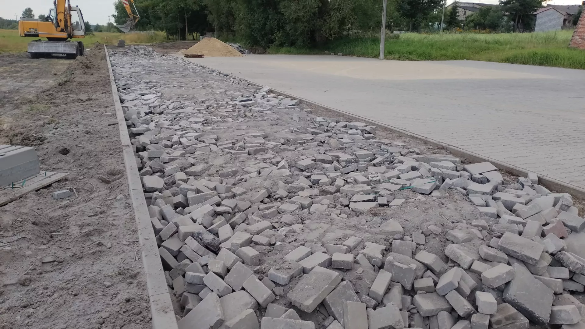 Rozszerzanie utwardzenia terenu w miejscu organizacji Jarmarku nad Liswartą. Widoczny ułożony krawężnik i wstępnie rozłożony gruz, a w tle także nawierzchnia wybrukowana kostką oraz pracująca koparko-ładowarka.