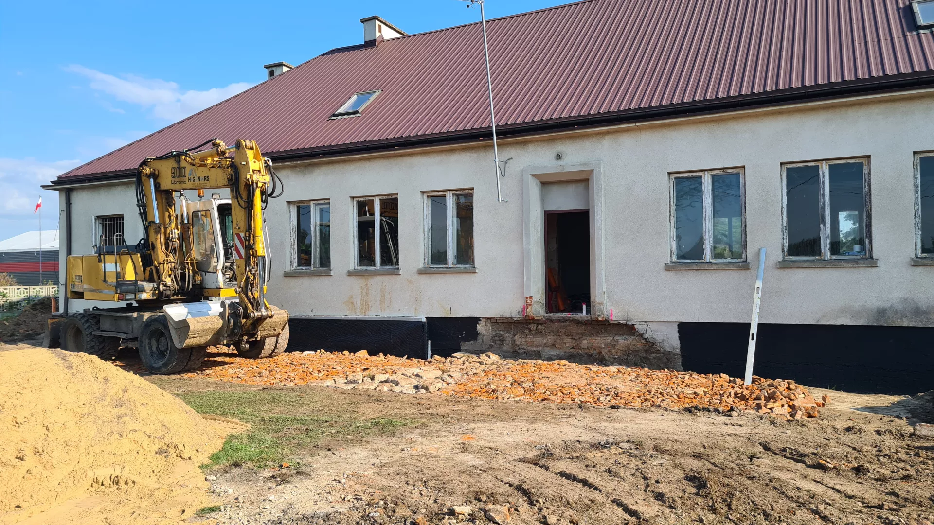 Prace remontowe przy budynku świetlicy w Grabarzach. Widoczna elewacja budynku i miejsce po zdemontowanych starych schodach wejściowych oraz teren utwardzony gruzem ceglanym. Obok po lewej pryzma piasku, a za nią koparko-ładowarka.