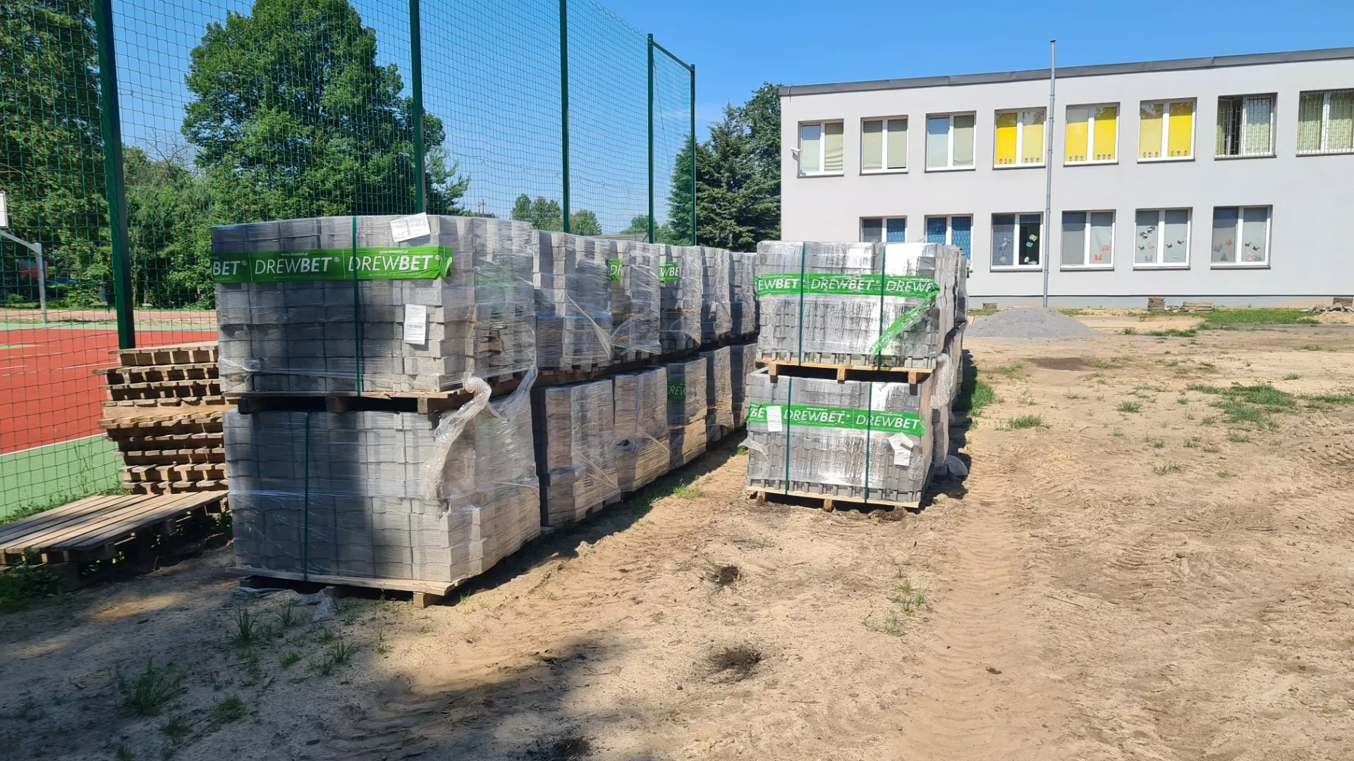 Kostka brukowa znajdująca się na paletach ustawionych przy ogrodzeniu boiska wielofunkcyjnego w Lindowie, w pobliżu widocznego w tle budynku szkoły.