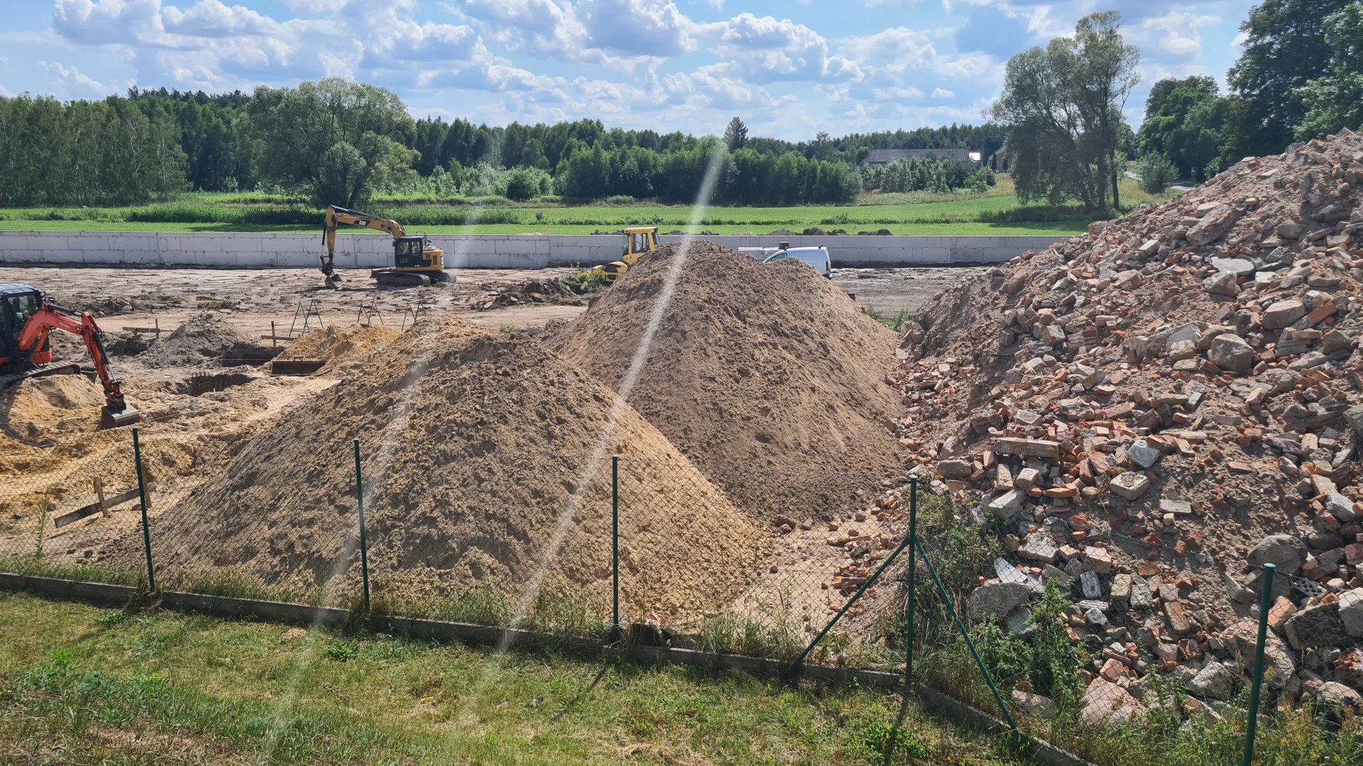 Pryzmy piasku i gruzu na ogrodzonym siatką terenie budowy PSZOK w Lipiu. NA dalszym planie widoczne koparki prowadzące roboty ziemne oraz jeden z wykonanych murów oporowych.