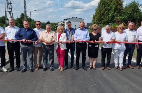 Uczestnicy ceremonii otwarcia drogi powiatowej przygotowują się do przecięcia biało-czerwonej wstęgi rozpostartej w poprzek nowej jezdni.