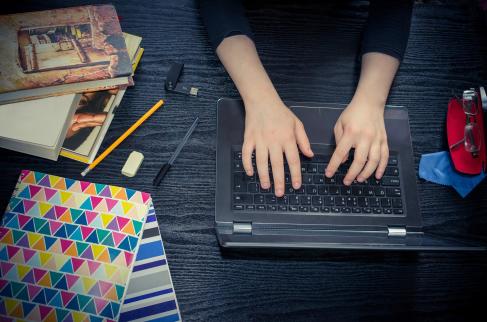 Ręce ucznia spoczywają na laptopie, który stoi na biurku w sąsiedztwie zeszytów, książek i przyborów do pisania.