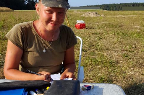 Studentka archeologii Paulina Tarka w warunkach polowych uzupełnia dokumentację badań, siedząc przy rozkładanym stoliku. Każde znalezisko i jego dokładne położenie podlegają skrupulatnemu dokumentowaniu.